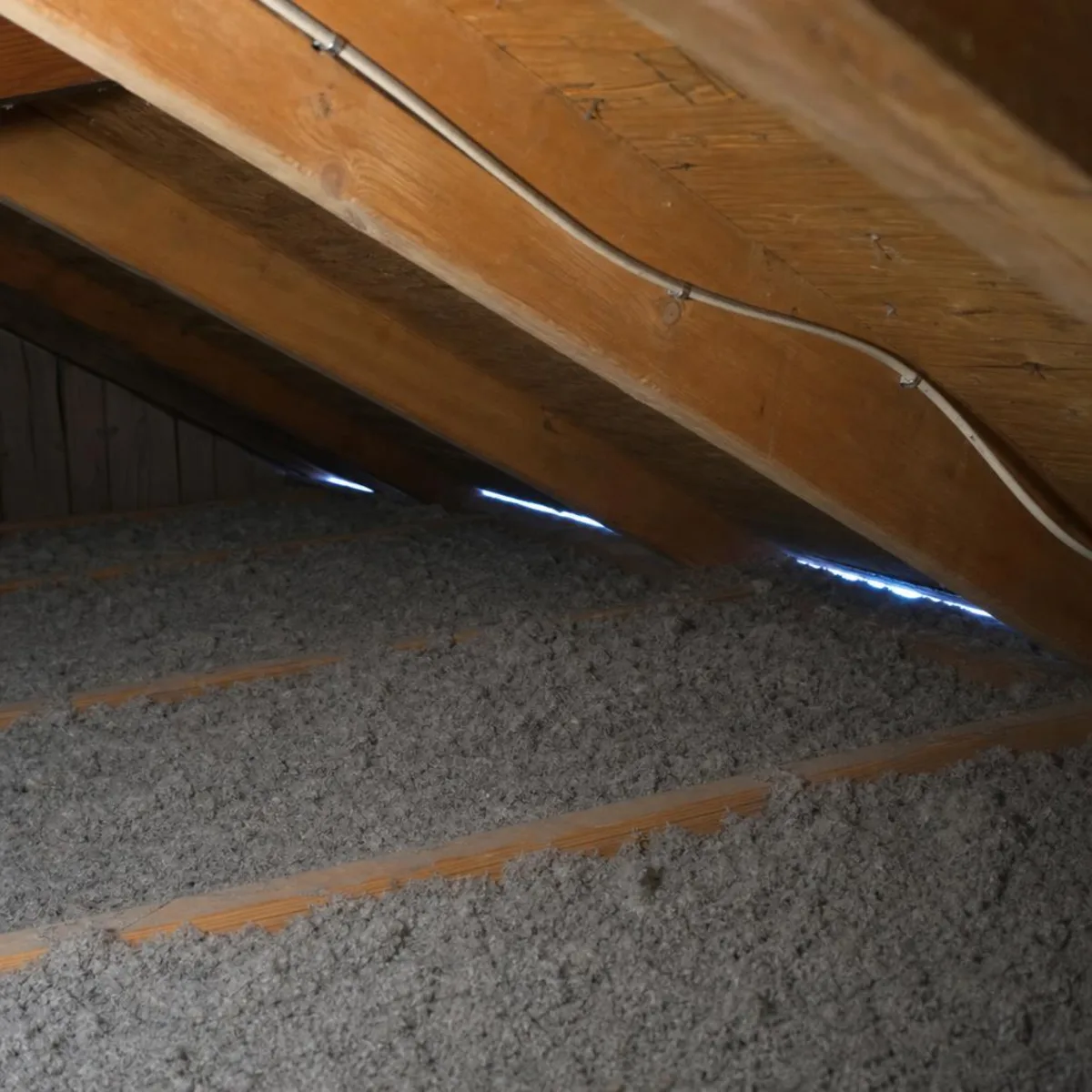An attic interior with blown-in insulation between the joists, electrical cable stapled along a rafter, and a thin line of daylight showing at the eave.