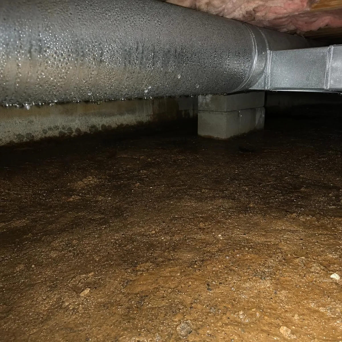 A metal duct running across a crawl space above dark, damp bare earth, with moisture staining in the soil beneath the run.