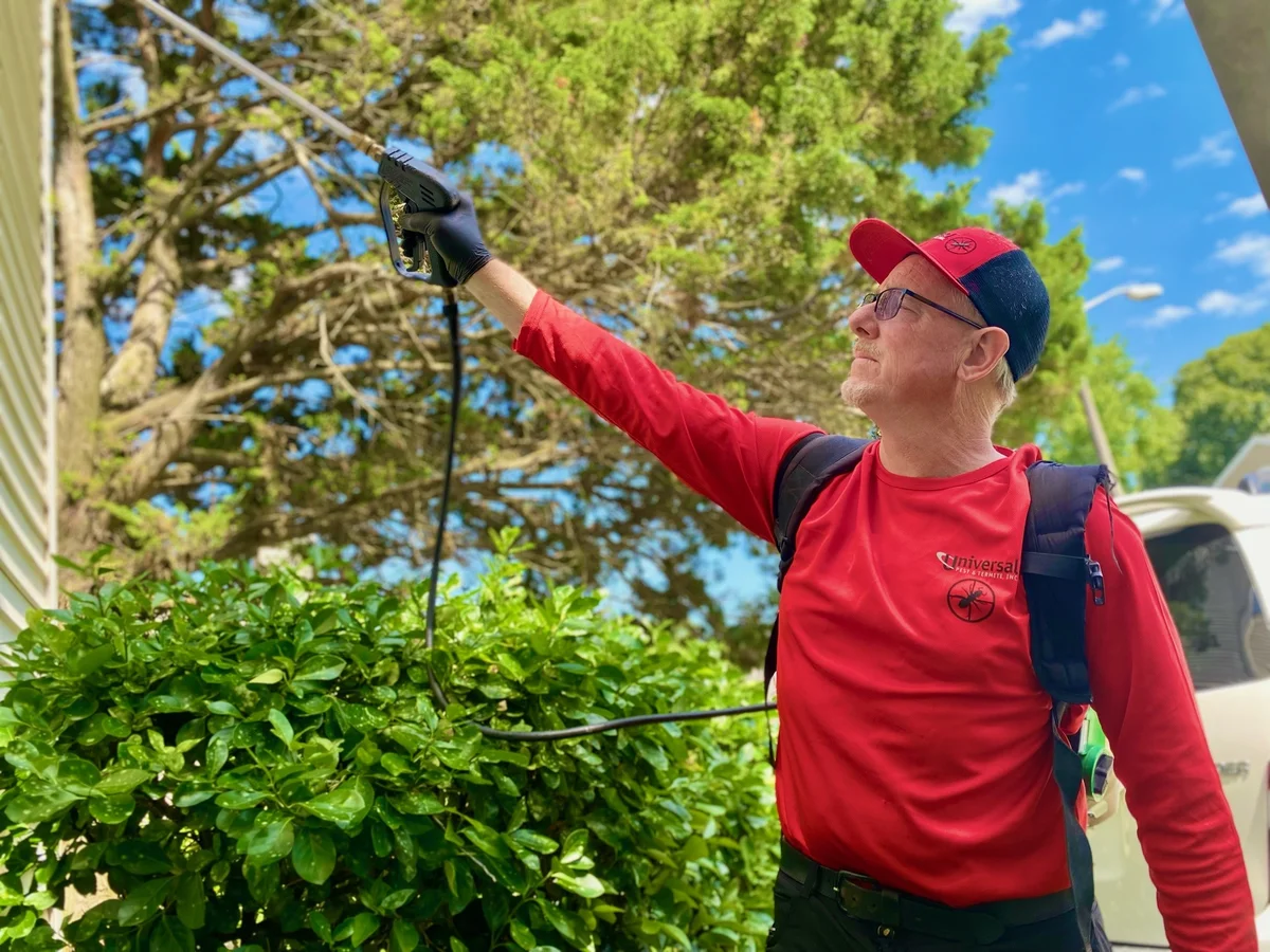 Universal Pest & Termite technician in a red shirt using a backpack sprayer to treat the eaves of a home next to the shrubs, with the company service van parked behind him